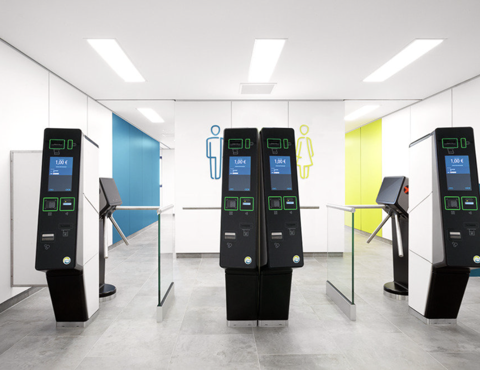 Moderne Sanitäranlagen-Zugangsschranke mit vier schwarzen Bezahlterminals in heller, sauberer Umgebung; im Hintergrund Symbole für Damen- und Herrentoilette